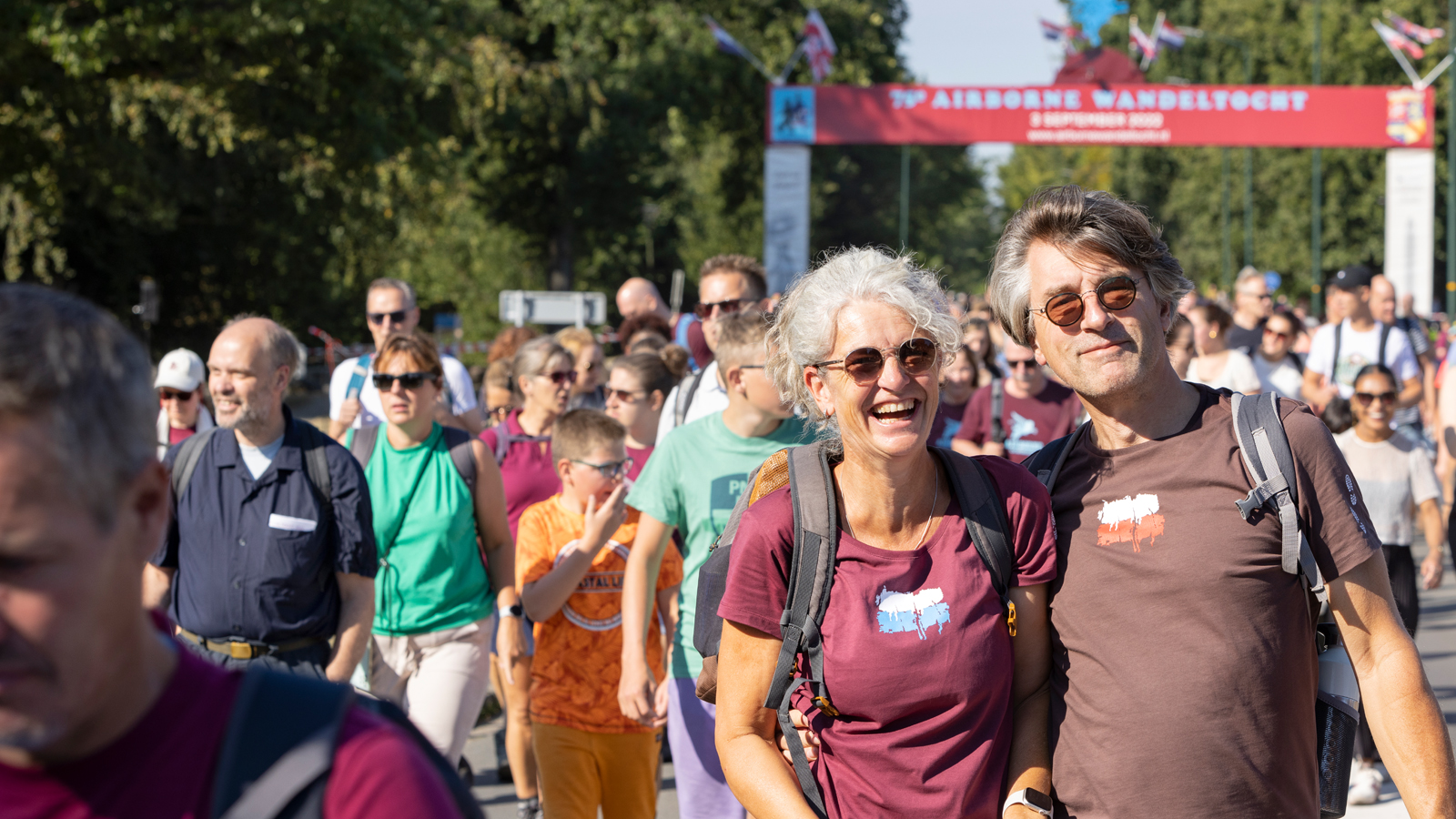 Een man en een vrouw kijken lachend in de camera terwijl er om hen heen mensen deelnemen aan een wandelevenement