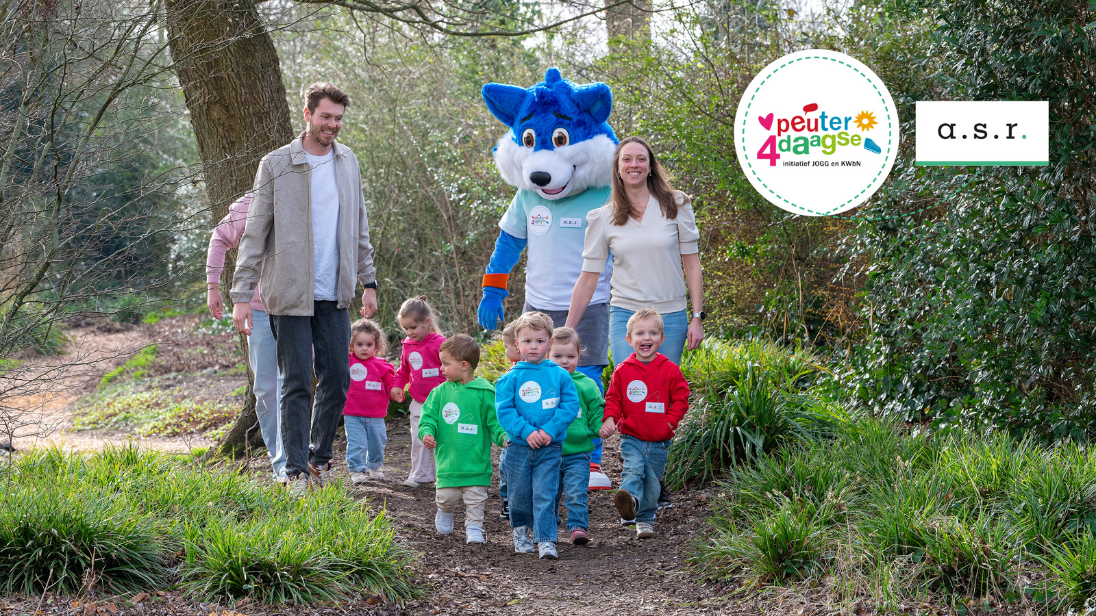 Volwassenen en peuters wandelen samen met mascotte Fiero over een bospaadje