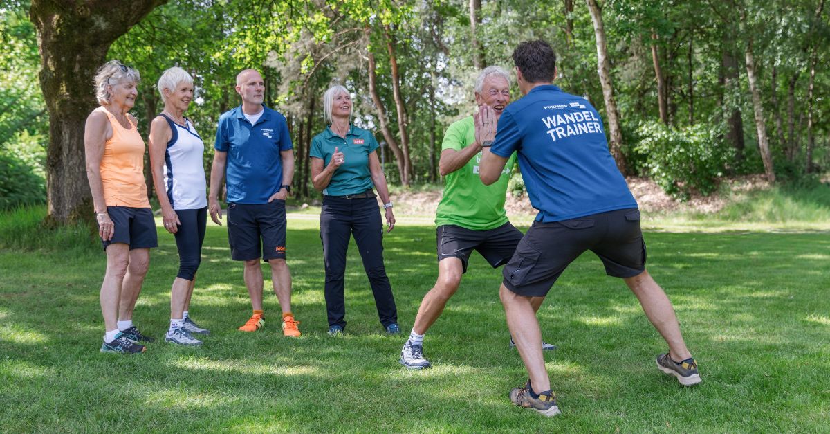 Twee wandeltrainers doen lachend een oefening, vier wandeltrainers kijken op de achtergrond toe