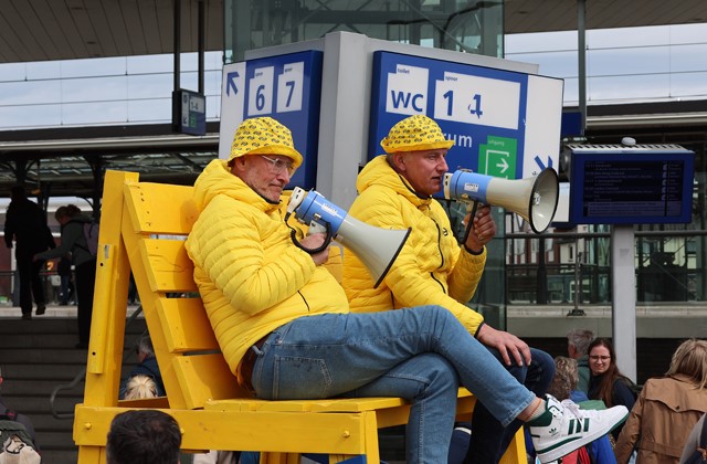 Twee mannen op een grote stoel in gele kleding met megafoons op een NS Station